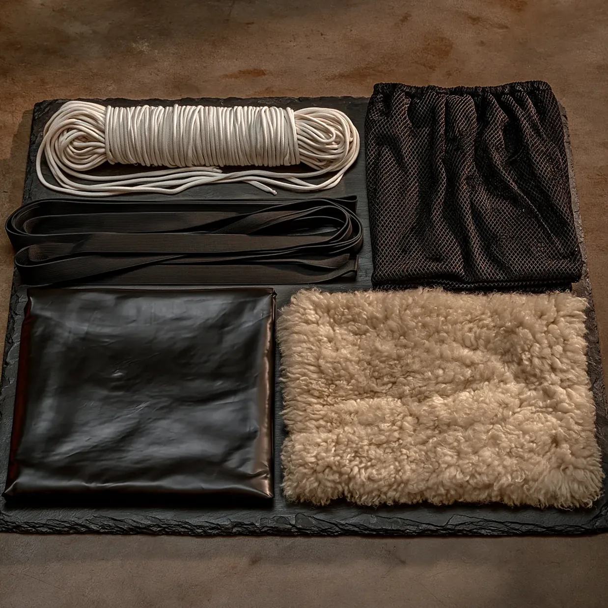 À plat sur une ardoise sombre : un cordon de coton blanc enroulé, des sangles élastiques noires, un carré de résille, une feuille de caoutchouc noir et un carré de peau lainée.