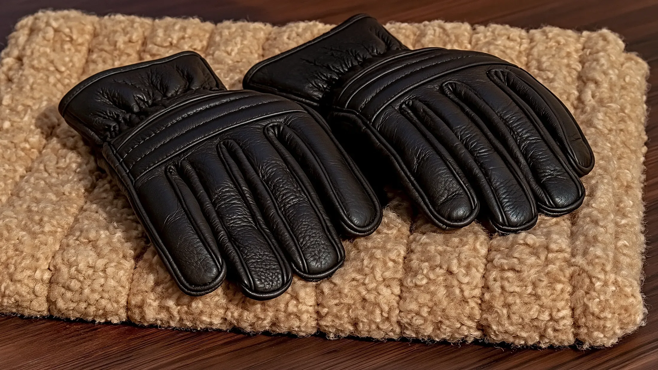 Deux gants de cuir noir posés sur un tapis de mouton crème, un petit poids de laiton à côté.